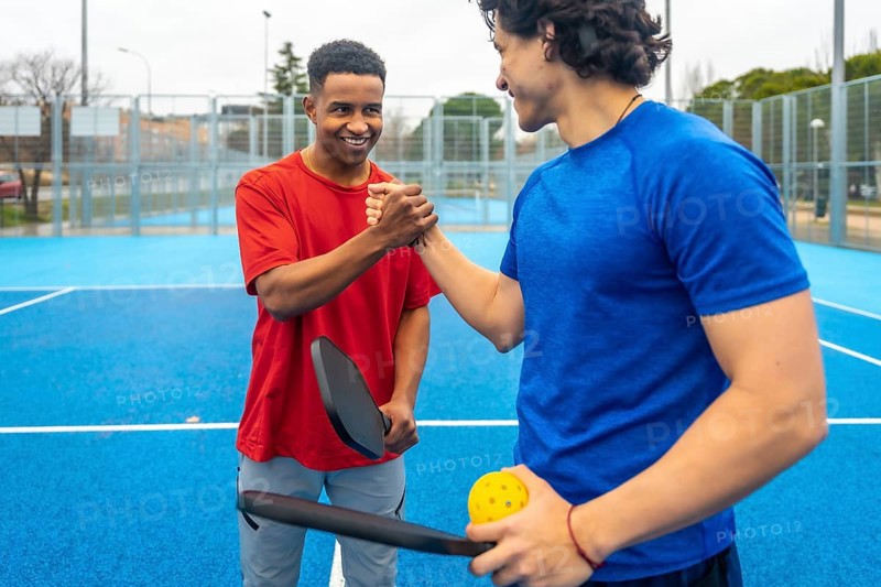 Two male pickleball players shaking hands on a blue outdoor court after a match, showing sportsmanship, friendship and teamwork in a casual, active urban summer setting