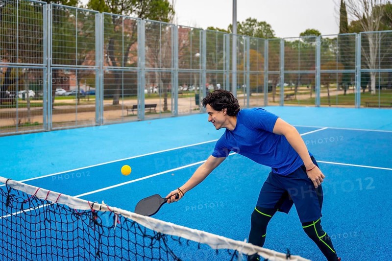 Young man in sports clothing enthusiastically playing pickleball on a bright blue court, hitting a yellow ball with a paddle, enjoying the active sport outdoors