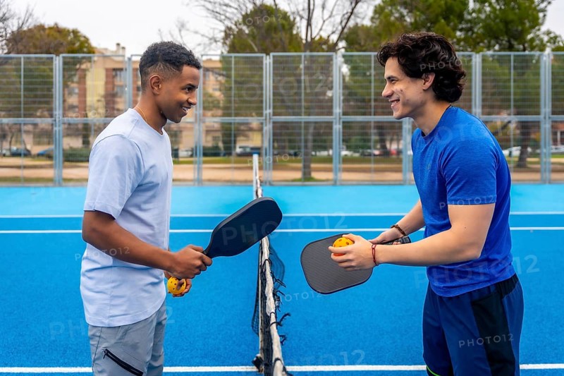Two diverse young men standing on a vibrant blue pickleball court, holding paddles and balls, enjoying a friendly game outdoors and sharing a moment of camaraderie and healthy competition