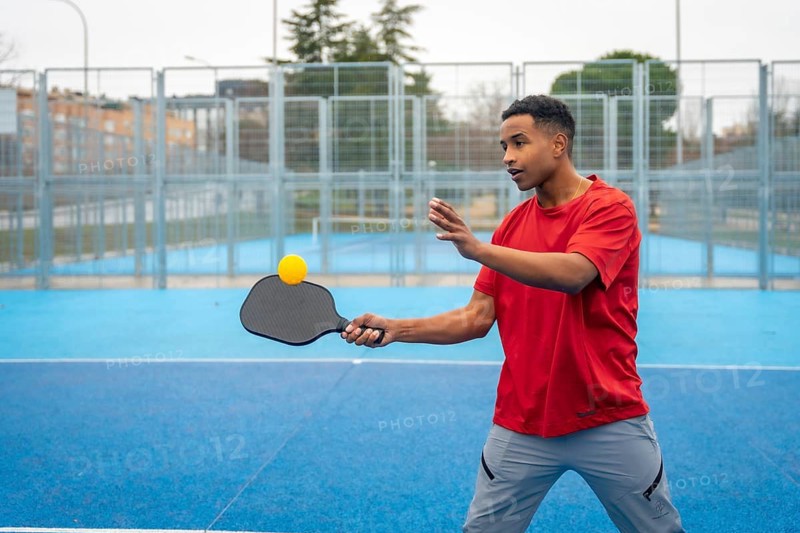 Young man concentrating on returning the ball during a pickleball game on a bright blue outdoor court, engaging in a healthy active lifestyle and recreational sport