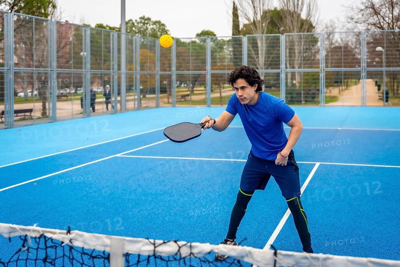 Young man in blue activewear playing pickleball on a blue court, intently hitting a yellow ball with a paddle, enjoying the modern racket sport outdoors