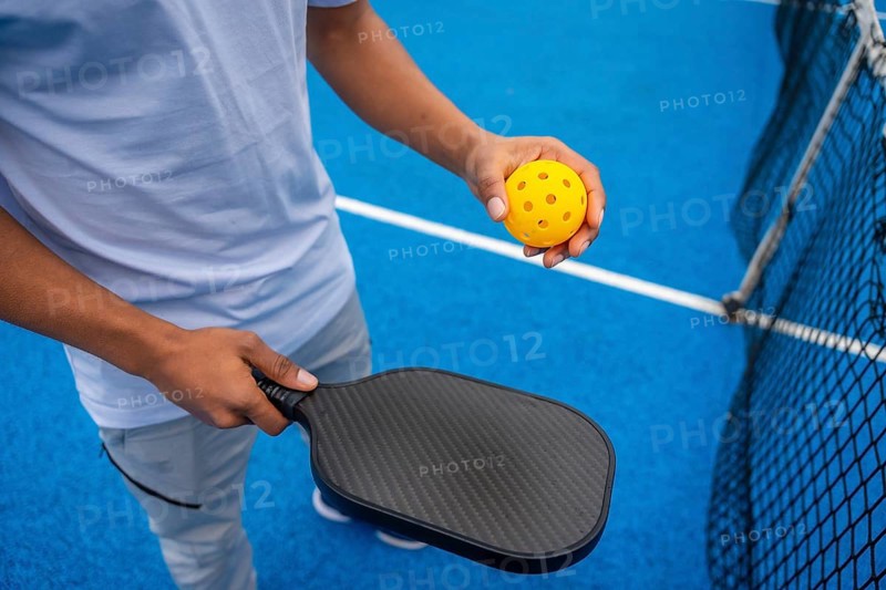Person standing on a bright blue pickleball court, holding a black paddle and a yellow perforated pickleball, ready to play and engage in competitive sport and active lifestyle