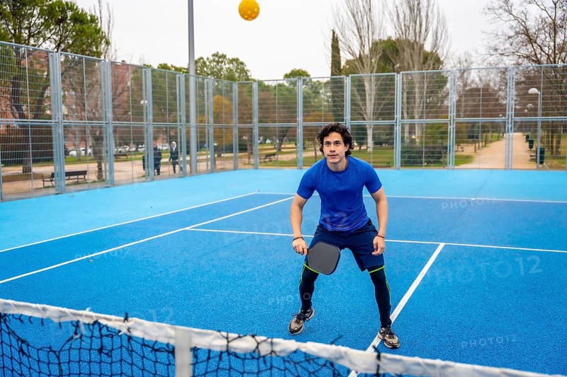 Young man actively playing pickleball on a vibrant blue outdoor court, holding a paddle and watching the ball during a game, portraying a healthy and active lifestyle