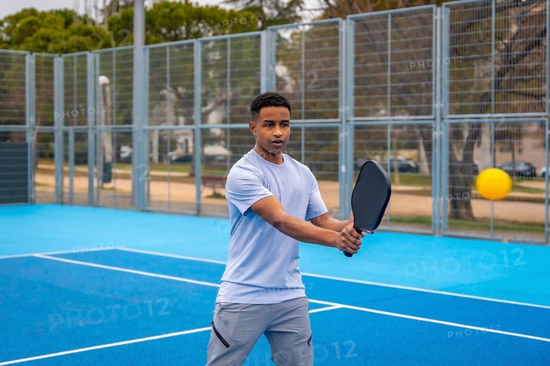 Young man poised to strike a yellow ball with a paddle on an outdoor blue pickleball court, focused and athletic during a competitive match on a sunny day