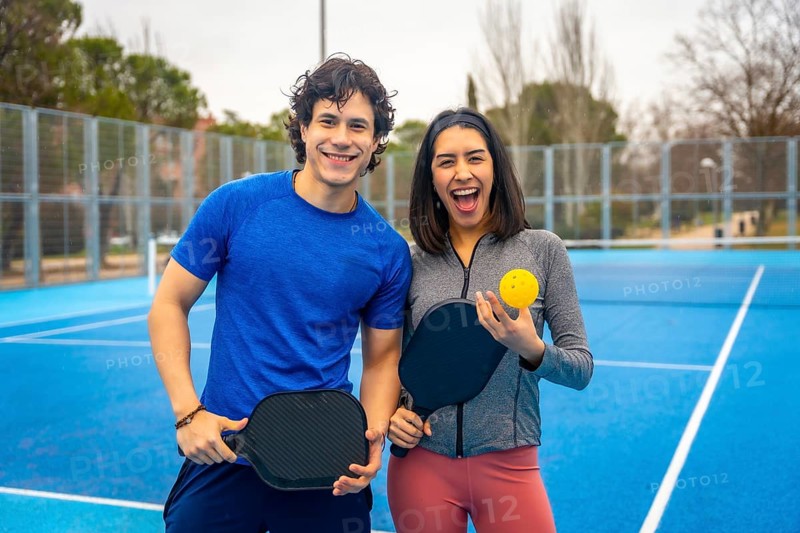 Two young athletes smiling on a bright blue outdoor pickleball court, holding paddles and a ball, standing together and ready to play a fun, active match outdoors