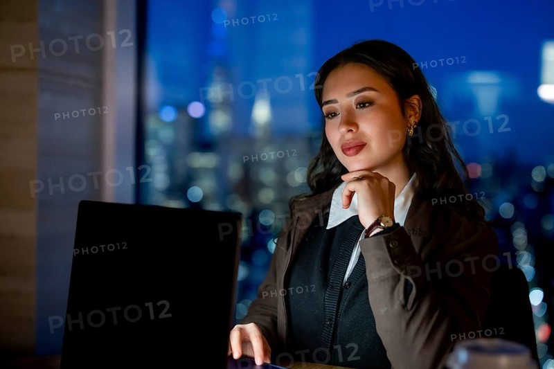 Young businesswoman with elegant hairstyle working late hours, researching or analyzing data on her laptop, overseeing the illuminated manhattan city skyline from her modern office
