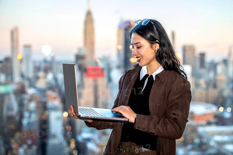 Young asian businesswoman using a laptop while standing against a blurred manhattan cityscape, representing urban professionalism, global business, and modern technology