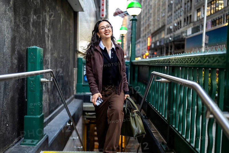 Young businesswoman smiling while emerging from a subway entrance in manhattan, carrying a smartphone and a handbag, navigating the urban environment of new york city during her daily commute