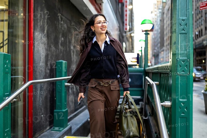 Young businesswoman with glasses and a professional bag emerging from a subway station entrance, confidently walking on a city street in new york, embodying urban success