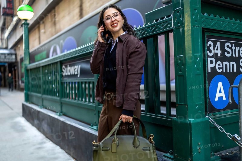 Young businesswoman smiling, talking on a mobile phone, and carrying a handbag while standing near a subway entrance in manhattan, new york, representing urban life and commuting