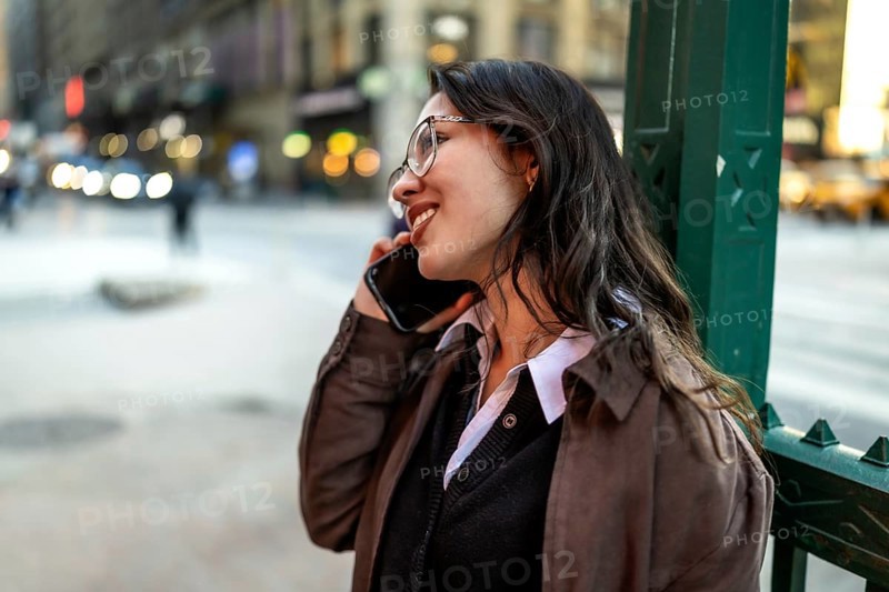 Young businesswoman smiling while talking on a smartphone, having a pleasant conversation standing in an urban setting with blurred city lights and vehicles in the background