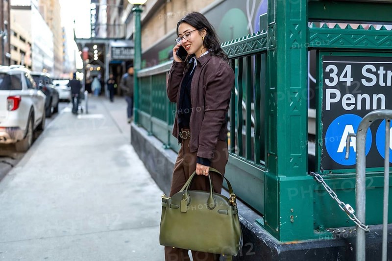 Young businesswoman walking on a new york city street, talking on her smartphone, carrying a stylish bag, with a subway station entrance visible in the background