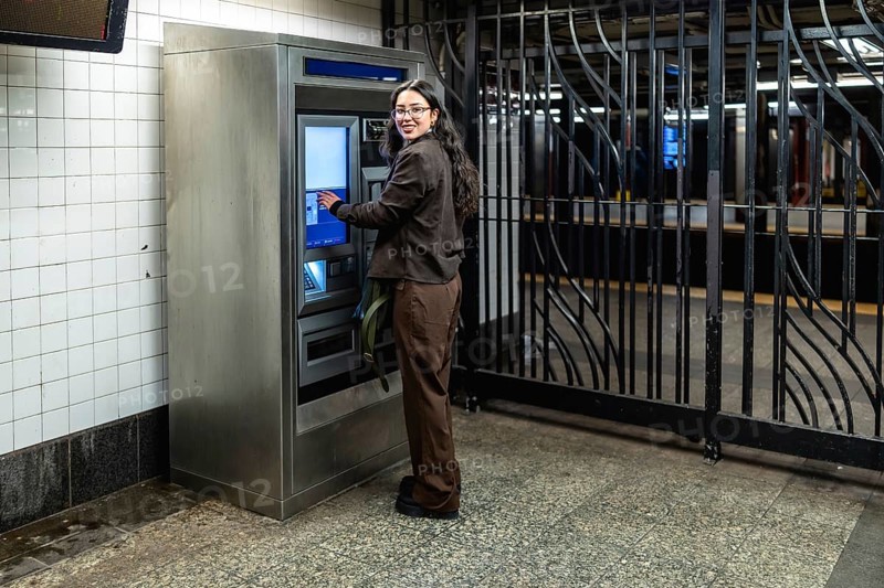 Young businesswoman with glasses and dark wavy hair smiling while interacting with a ticket machine at a subway station, preparing for her daily commute in manhattan