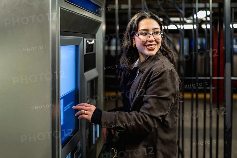 Young businesswoman with glasses and dark coat smiling, looking over her shoulder while interacting with a subway ticket machine in a busy urban station