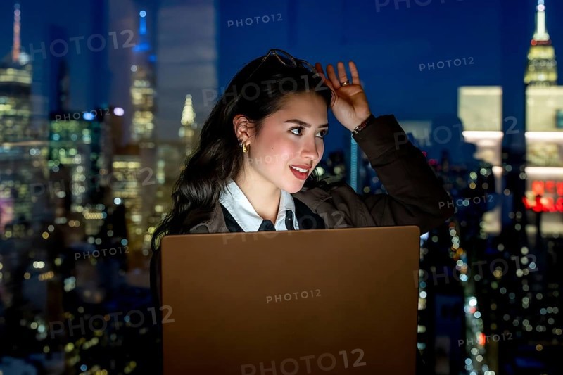 Young businesswoman working on laptop, thinking and looking away from a high rise office window at the illuminated manhattan skyline and city lights at night