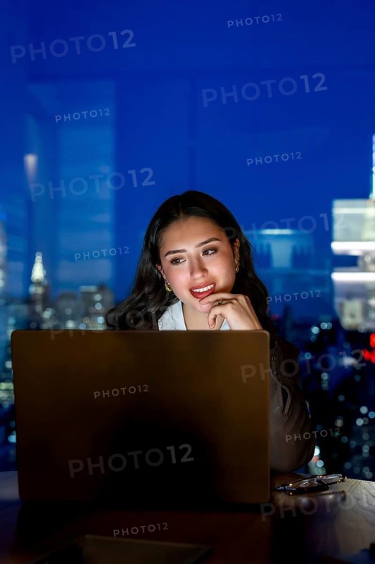 Young businesswoman working late on a laptop inside a modern manhattan office, illuminated by the screen light with the urban skyline of new york city visible through the window at night