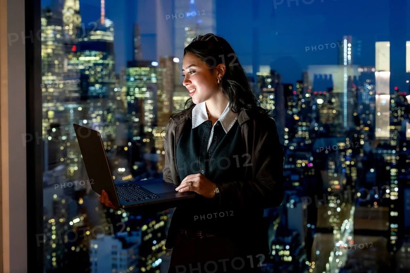 Young businesswoman stands by a floor to ceiling window at night, holding a laptop and smiling at manhattans illuminated skyline, symbolizing ambition, leadership and modern work life