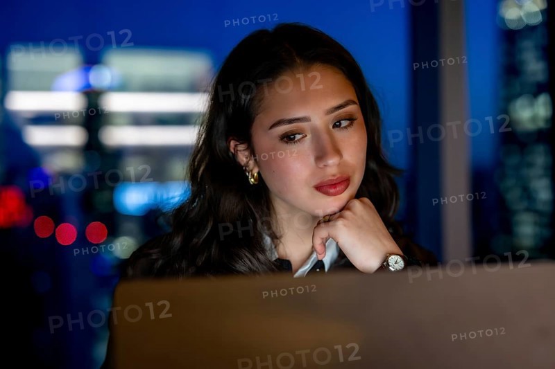 Young businesswoman working on a laptop at night, focused on her tasks in an office with the illuminated manhattan city skyline visible through the window, symbolizing dedication and ambition
