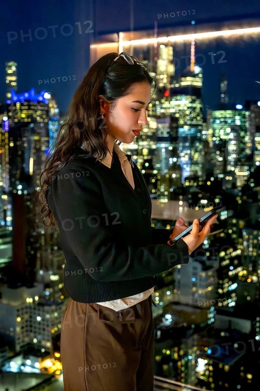 Young businesswoman standing by a window, engrossed in her smartphone, with the illuminated manhattan skyline serving as a dynamic backdrop, symbolizing connectivity and urban professional life