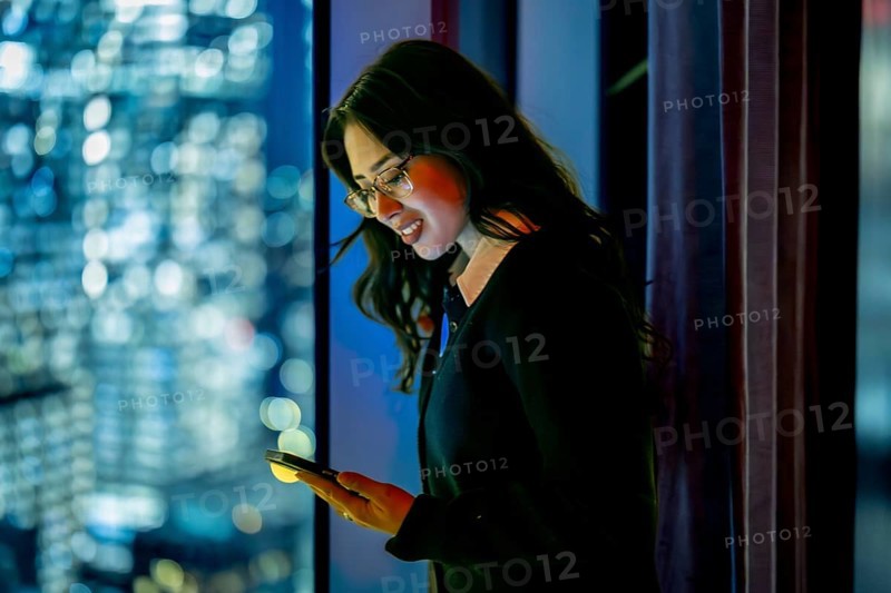 Young businesswoman standing by a skyscraper window at night, focusing on her mobile phone screen bathed in its glow, representing global communication and urban success