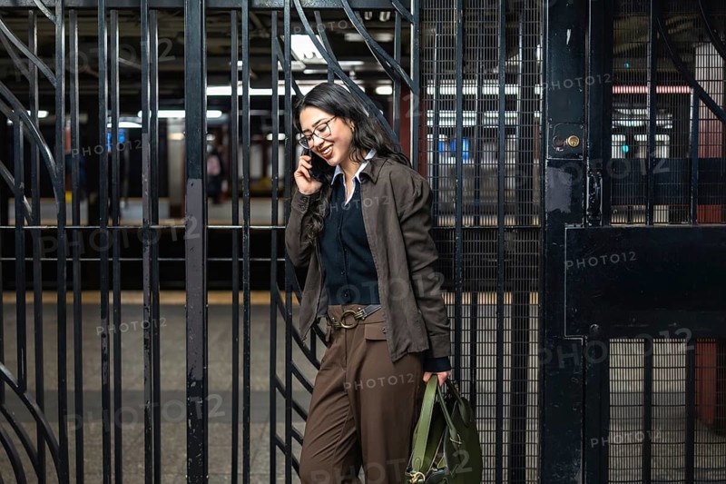 Young businesswoman standing by a subway gate, holding a bag, and smiling while having a phone conversation, representing urban professional life and commuting
