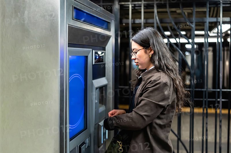 Young businesswoman with glasses and sleek dark hair interacting with a subway ticket machine, managing her urban commute and digital transaction in a bustling city environment