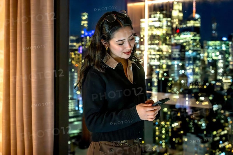 Young businesswoman standing by a window, focused on her smartphone while managing business communications with the illuminated manhattan skyline in the evening