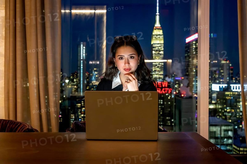 Young businesswoman with glasses on her head, focused on her laptop in a high rise at night, overlooking the illuminated new york city skyline and empire state building