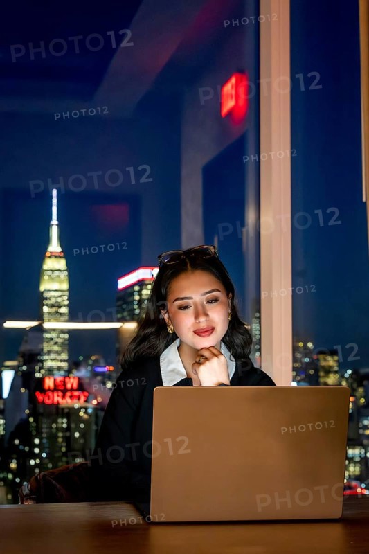 Young asian businesswoman focusing on her laptop screen, working late from an office with the illuminated manhattan skyline, including the iconic empire state building, visible outside the window