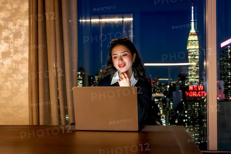Young businesswoman focusing on her laptop screen, working late in a high rise office or hotel room overlooking the illuminated new york city skyline and the iconic empire state building at night