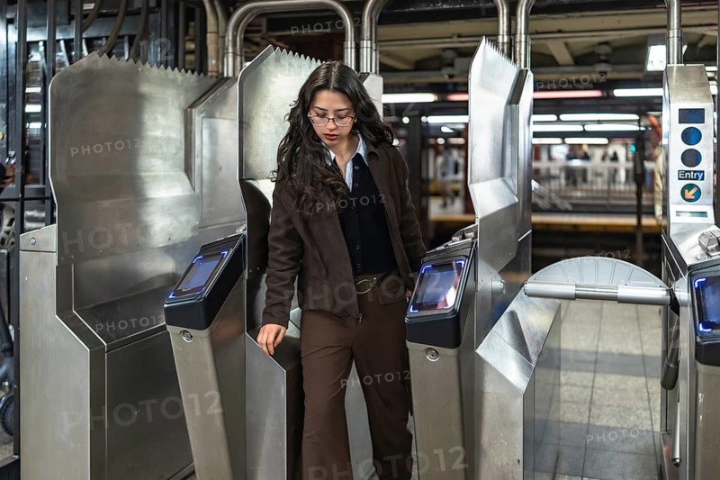 Young professional woman with glasses and dark wavy hair passes a modern subway turnstile in manhattan, commuting through an urban underground station during rush hour