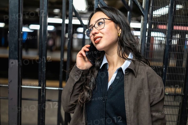 Young businesswoman in glasses talks on smartphone while waiting for a manhattan subway, multitasking and embodying urban connectivity and a professional on the go lifestyle