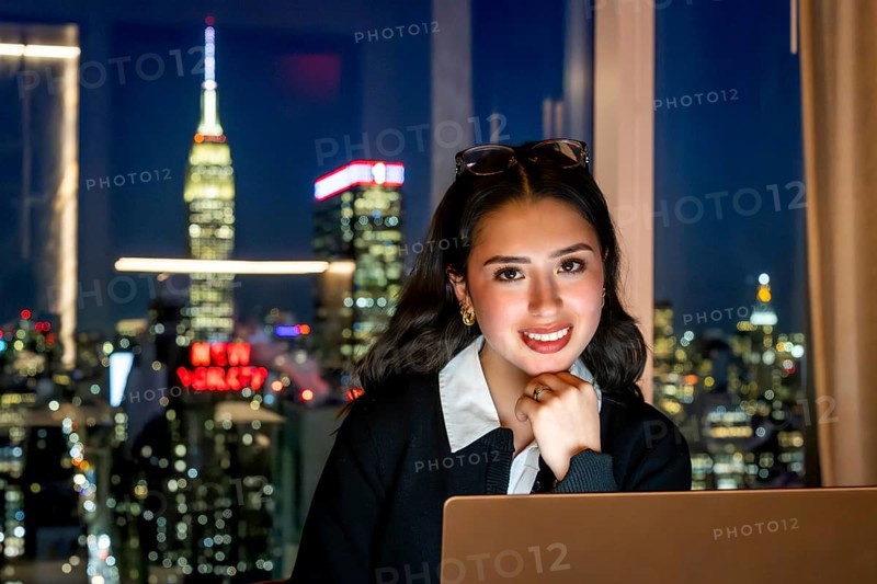 Young businesswoman smiling at camera, sitting in front of a window with a blurred night view of the illuminated new york city skyline and the empire state building