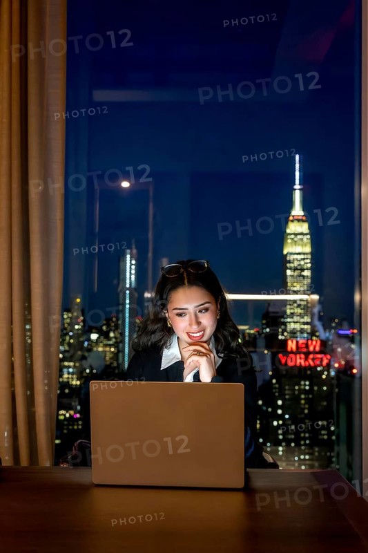 Young businesswoman observing laptop screen with a thoughtful smile, working late in a high rise office overlooking the illuminated manhattan cityscape and iconic buildings