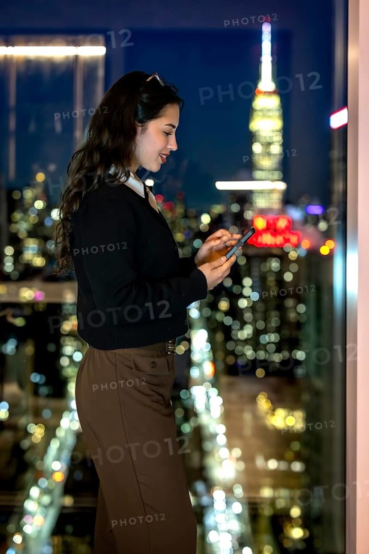 Young executive girl texting or browsing on a smartphone, standing by a window with a blurred view of new york city, the empire state building glowing under the dark night sky