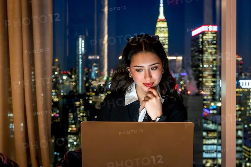 Young businesswoman working on her laptop with a contemplative expression, overlooking the illuminated manhattan cityscape from a high rise office at night