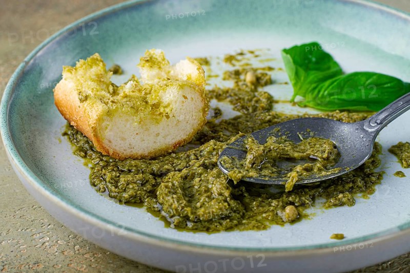 On a platter is a pesto sauce made from fresh basil, with a slice of bread and a spoon on a plate. The pesto sauce looks creamy and green, and the bread is partially eaten
