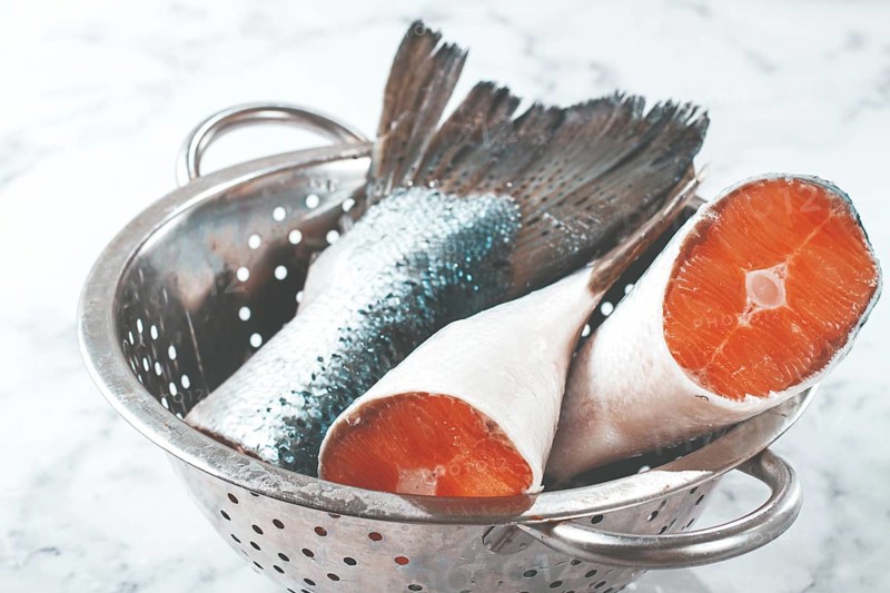 Fish tails are placed in a colander on a marble counter. The colander allows water to drain away from the fish. The fish looks fresh and clean, ready for cooking or preparation