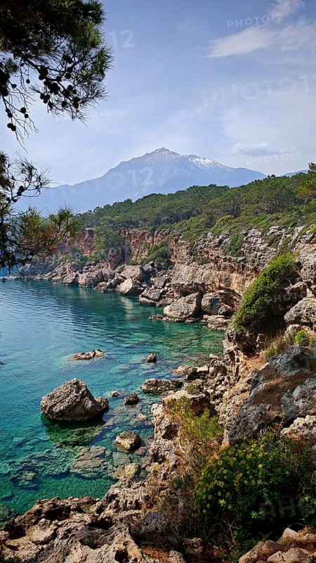 Rocky coast with clear blue water and view of snow-capped mountains in the background, view of Tahtali on the Lycian Way, Antalya, Lycia, Turkey
