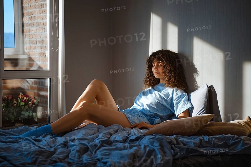 Young woman sits on a bed in a room with sunlight streaming in from a window. She has curly hair and wears a light blue pajamas and socks
