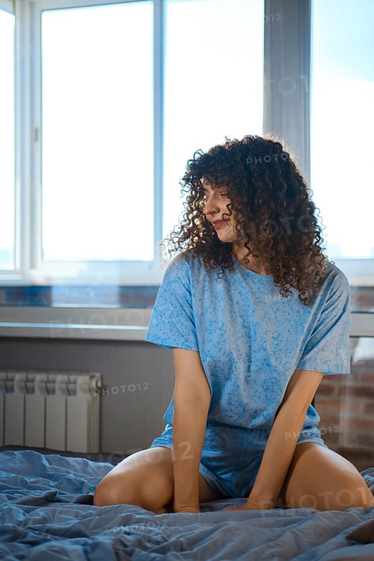 A young woman is sitting on a bed in a simple room. Bright daylight comes through the window, highlighting her curly hair and relaxed pose. The bedding looks soft, and the walls have a warm tone
