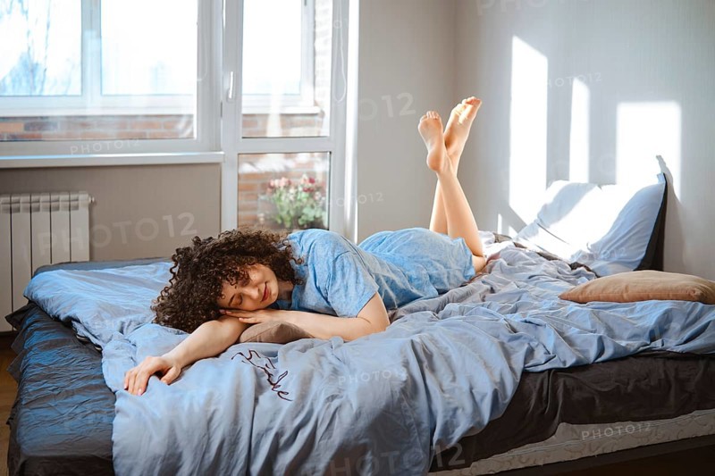 A young woman lies on her stomach on a bed, wearing blue pajamas. Sunlight comes through the window and shine her feet. Plants are visible outside. The room feels bright and airy