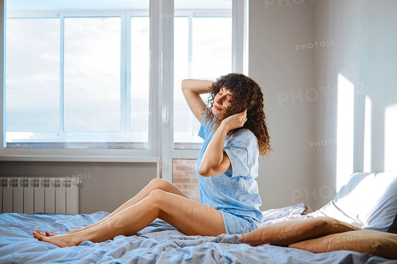 A woman sits on her bed in a bright room. She stretches her arms and turns her head as sunlight comes through the window. The room has a cozy feel with soft bedding and pillows