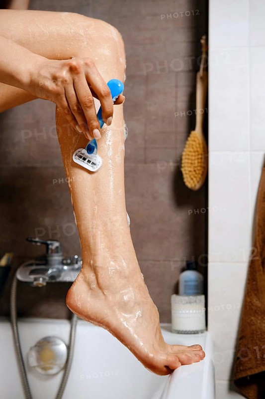 Unrecognizable woman put her leg on the edge of a bathtub and using a razor to shave her leg. Shaving cream covers the skin. The bathroom has a tidy setup with bath products