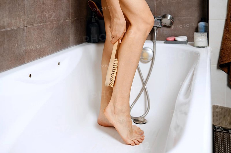 Woman stands in a bathtub during a personal care session. She hold a wooden massage brush in one hand and use it on her leg. Items are placed nearby for convenience