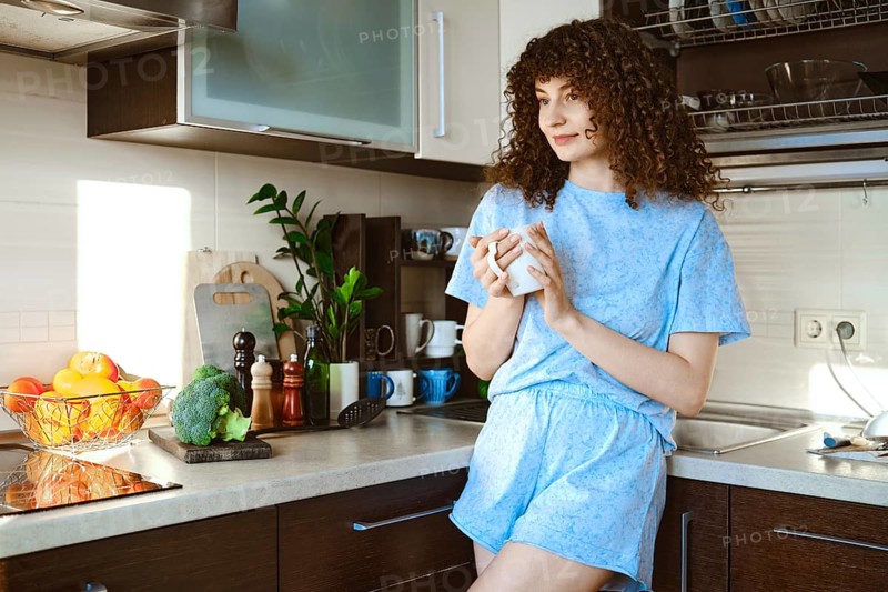 Smiling woman stands in a kitchen, sipping coffee from a mug. Fresh fruits and vegetables are on the counter. Bright light comes through the window, creating a warm atmosphere in the room
