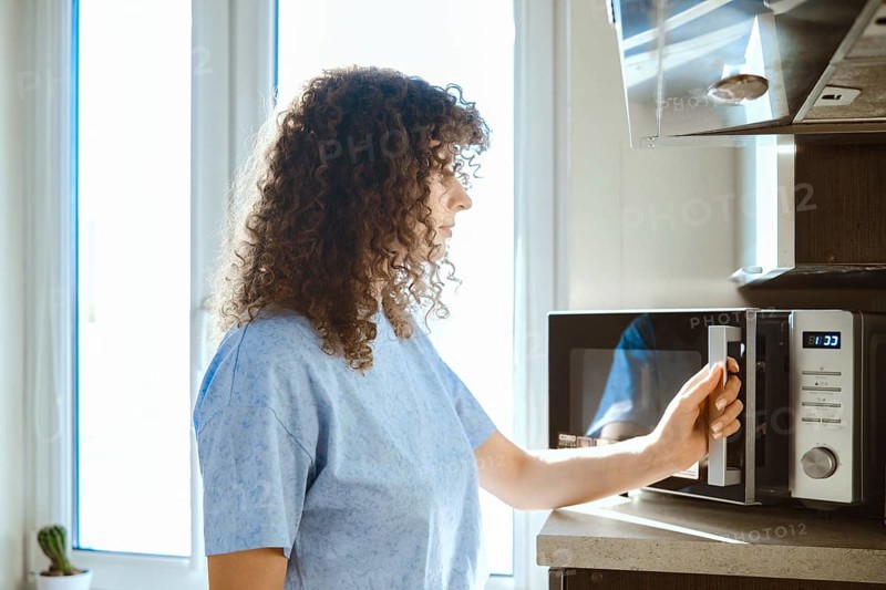 A woman stands by a microwave in a kitchen. She is preparing to heat food. Sunlight enters through the window. The kitchen is tidy with simple decor. There is a plant in the background