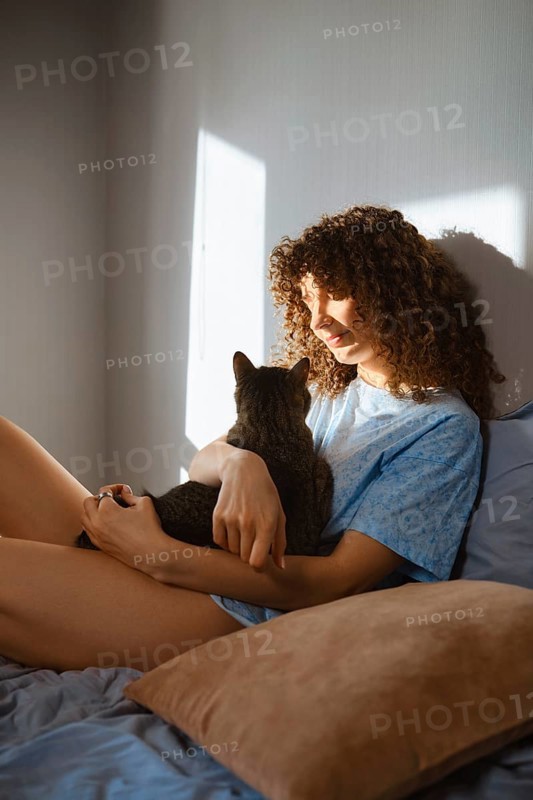 A woman with curly hair sits on a bed while holding a cat. Sunlight comes through the window, creating a warm atmosphere in the room. The moment shows a connection between the woman and her pet