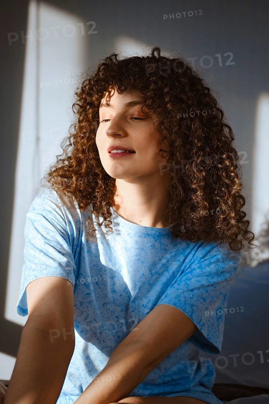 A woman with curly hair sits in a well-lit room. Sunlight streams through the window, creating warm shadows. She looks relaxed and happy, enjoying her moment in the soft light of the morning