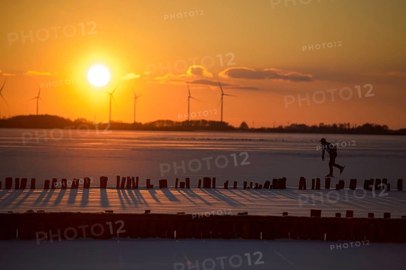 Ice skaters in front of sunset over icy landscape with wind turbines and intense colors on the frozen Dümmer Lake, Lembruch, Diepholz district, Lower Saxony, Germany